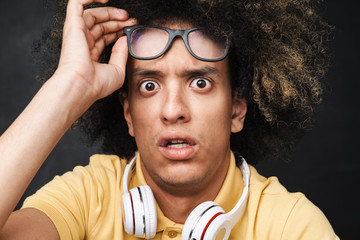 Scared teenage guy posing over grey chalkboard wall with headphones wearing eyeglasses.