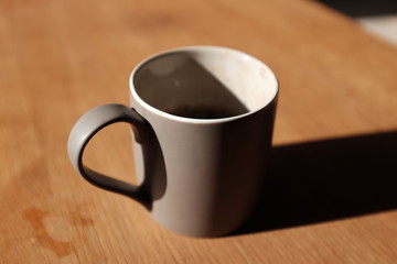 A half-finished coffee drink in a grey mug, casting a long shadow onto a coffee table one summer’s morning.