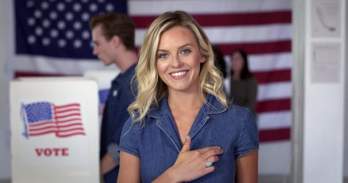 MCU Young blonde Caucasian woman in denim dress puts on "I Voted" sticker and smiles, standing proud in front of polling booths with US flag. Fluid head tripod, real-time 4K