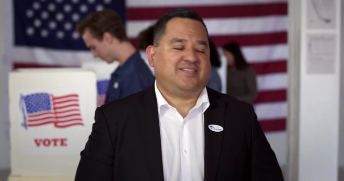 MCU Hispanic man in white shirt and black jacket puts on "I Voted" sticker and smiles, standing proud in front of polling booths with US flag. Fluid head tripod, real-time 4K