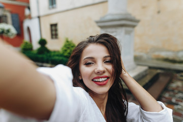 Close up horizontal pretty happy lovely smiling young woman making selphie with mobile phone on the background of street and fixing her hair.