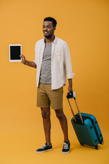 happy african american man standing with luggage and holding digital tablet with blank screen on orange