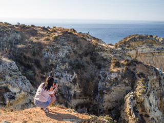 Young lady taking a photo of the colourful cliffs of Ponta da Piedade in Algarve. Mornuing (low) sun illuminated, lady and cliffs in foreground, ocean and sky in background