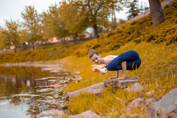 Slender young brunette yogi performs challenging yoga exercises on the green grass in autumn against the background of nature