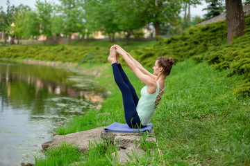 Thin brunette girl sitting in Both big toe exercise, Paripurna Navasana pose  in a summer park. Green forest on the background. Woman doing exercises on the yoga mat