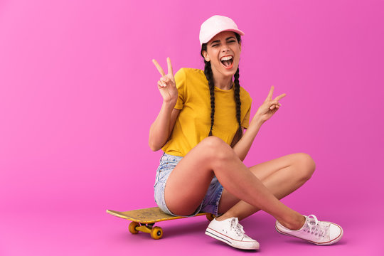 Image of smiling happy woman wearing casual clothes showing peace sign and sitting on skateboard