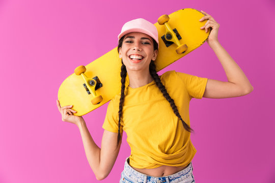 Image of pretty teenage woman wearing cap laughing and holding skateboard on her shoulders