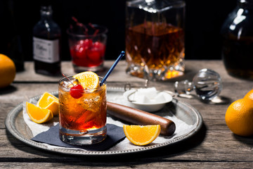 Old Fashioned Cocktail On Ice with Cherry and Orange Garnish and Ingredients on Bar in Dark Background