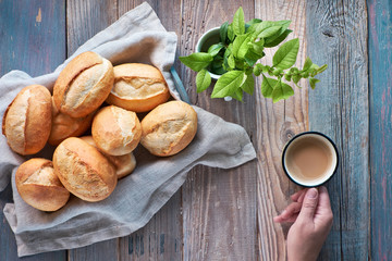 Bread buns in basket on rustic wood, spring leaves and hand with cup of coffee
