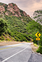 Two lane paved road with road sign indicating Winding Road Ahead