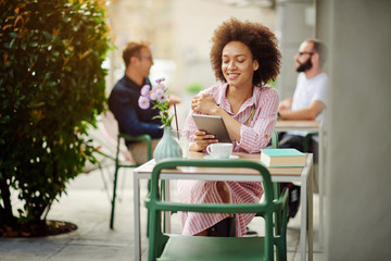 Smiling grogeous mixed race woman in pink striped dress sitting in cafe and using tablet. On table are cup of coffee, book and lemonade.