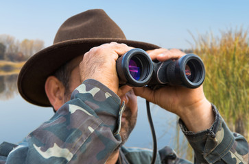 A hunter in a hat with binoculars looks out for prey against the background of the sunset.	