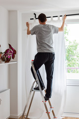 Man stands on a stepladder near the window and hangs white curtains on the curtain rod