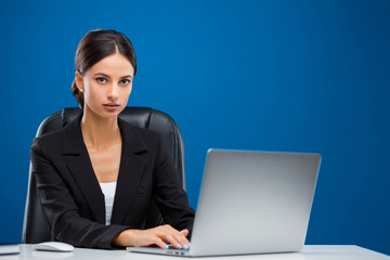 Young businesswoman sitting at her desk working at her laptop while looking at the camera, isolated on blue background.