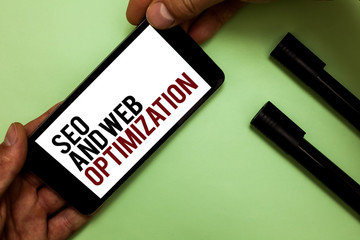 Text sign showing Seo And Web Optimization. Conceptual photo Search Engine Keywording Marketing Strategies Man's hand hold phone with black and red words near two black marker