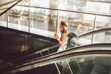 Portrait Smiling Woman Wearing Hat In Airport At Escalator. people traveling with hand luggage. Theme tourism and transport. Caucasian girl with coffee in airport terminal on an escalator