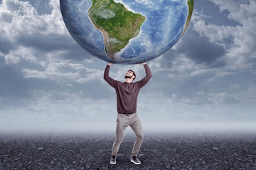 Full length front view of young man in casual clothes holding up big Earth globe on gloomy overcast day.
