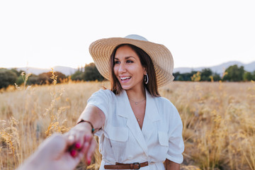 portrait of young beautiful caucasian woman outdoors at sunset in a yellow field. wearing a modern hat and smiling. Holding hands, follow me concept