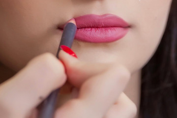 Fototapeta premium The female hand of visagiste is holding a pencil and drawing lips . Professional Makeup artist applies pencil lipstick bridal . Close up of unrecognizable pretty girl getting her lips painted.