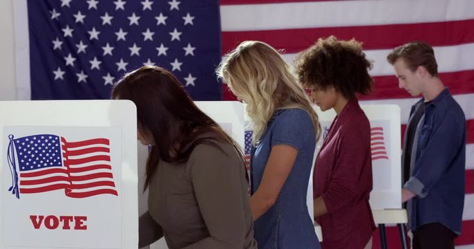 MS Series of voters of various demographics leaving and arriving at voting booths in polling station with US flag. Locked off continuous shot, real time 4K