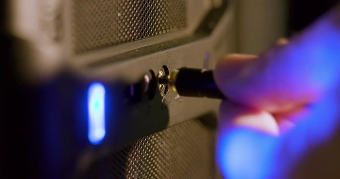 A blue light from a computer that indicates it is on reflects off the hand of a Caucasian male that is plugging a headphone jack into the designated socket. Macro Shot, Extreme shallow DOF
