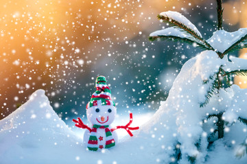 Small funny toy baby snowman in knitted hat and scarf in deep snow outdoors on bright blue and white copy space background. Happy New Year and Merry Christmas greeting card.