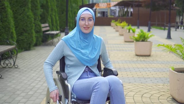 Portrait smiling young muslim woman disabled in traditional scarf sitting in a wheelchair in the park
