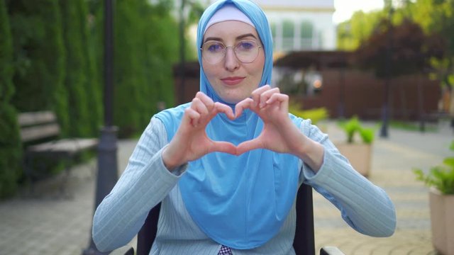 Portrait smiling young muslim woman disabled in traditional scarf sitting in a wheelchair in the park and shows hands heart shape