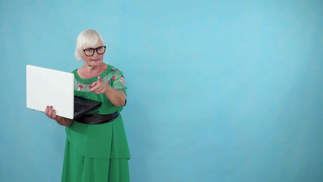 thoughtful old woman with a laptop in her hands finds a solution and stands on a blue background copy space