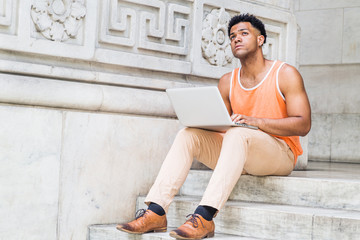 Fototapeta na wymiar Young Mix-Race American college student studying in New York City, wearing orange tank top, beige pants, brown leather shoes, sitting on stairs outside office on campus, working on laptop computer..