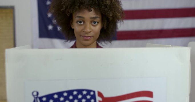 Close up, camera moves from voting booth up to young mixed race woman casting vote at polling station. She looks up to smile at camera. Large US flag on wall behind. Hand held, slow motion 4K 60fps