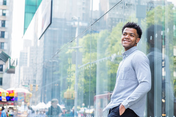 Fototapeta premium Young Mix Race American Man wearing light gray, long sleeve shirt, black pants, hand in pocket, standing against glass wall with reflections on street in Midtown of Manhattan, New York City, smiling..