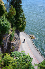 Cyclist rides on Stanley Park's famous Seawall on a sunny summer day. scenic view from the rock of water and majestic trees - obrazy, fototapety, plakaty