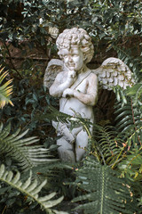 Closed up vintage style angel stone sculpture in the garden, with blurred green plants in background,little cupid angel with spread-ed wings