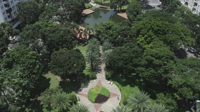 long wide aerial view of modern urban park featuring lakes, lush gardens, bridges, green space, pathways and trees. Also there are are buildings offering shade from the tropical sun and rain.