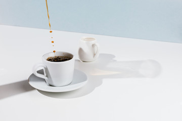 Closeup of cup with pouring coffee, saucer and creamer on white table