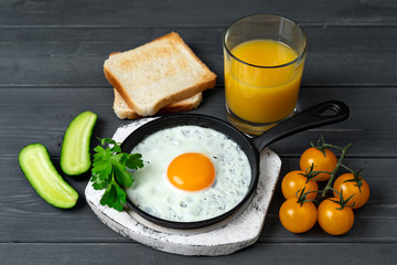 fried eggs in cast iron pan on white wooden stand with yellow cherry tomatoes, orange juice, toasts and cucumbers.