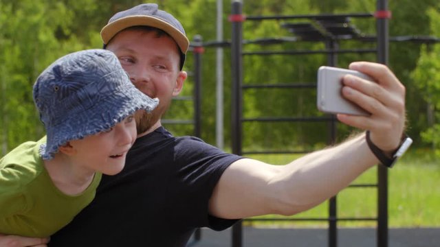 Waist-up shot of sweaty Caucasian father holding toddler son in sun hat and taking selfies together on smartphone after workout at outdoor gym in park, looking at camera, smiling and laughing