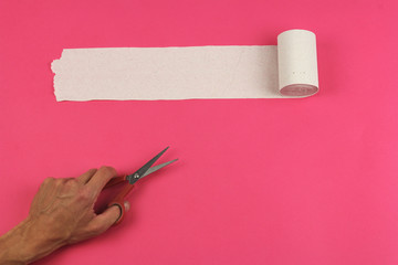 unrolled roll of rough recycled white toilet paper on pink background and human hand with scissors,...