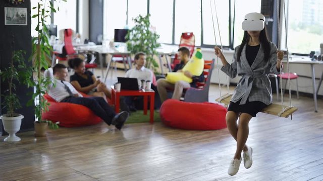 Young asian girl in futuristic white virtual reality glasses riding on a swing, modern office and workers on a blurry backround.