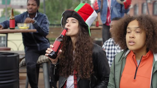 Young caucasian woman drinking beer, cheering for favorite team and then yelling in excitement while watching soccer match on TV in outdoor bar with african american female friend