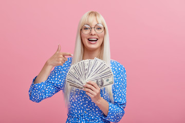 Portrait of a cheerful young blond woman in glasses  holding money banknotes and celebrating isolated over pink background