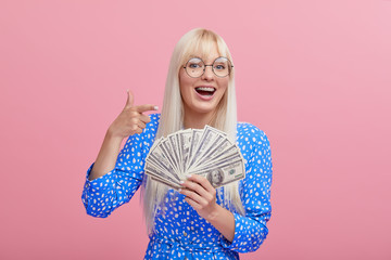 Portrait of a cheerful young blond woman in glasses  holding money banknotes and celebrating isolated over pink background