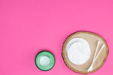 Cotton pads and sticks on round wooden stand, white cream in green glass jar, bright pink background. Cosmetology, healthcare, make-up concept. Top view, flat lay, copy space, mock up, layout design.
