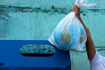 Hand putting Earth globe in trash bin. Planet in plastic bag on old turquoise concrete wall background. Environmental pollution idea. Ecology protection, zero waste, no plastic concept