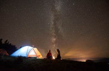 Night summer camping in the mountains. Romantic couple hikers having a rest together, sitting beside campfire and glowing tourist tent under beautiful night starry sky full of stars and Milky way.