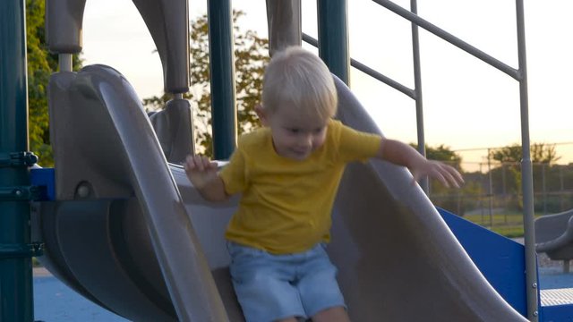 Little boy (toddler, two years old kid) having fun, sliding down. Outdoor plastic playground slide for children activities in park. Summer time, sunset, backlight, golden hour