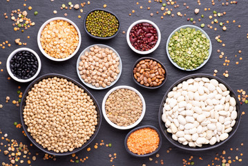 Legumes, lentils, chickpeas and beans assortment in various bowls on black stone background, top view
