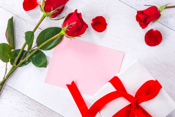 blank pink card decorated with gift box and roses on white wooden table, top view