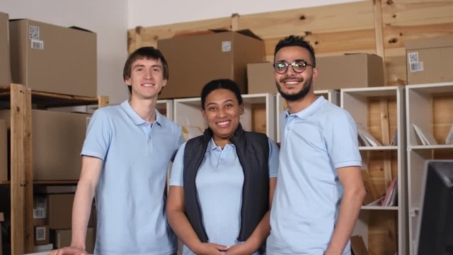 Thigh-up shot of ethnically diverse male and female post office clerks posing together during work shift, wearing uniform polo shirts and standing in front of storage shelves with parcels and mail